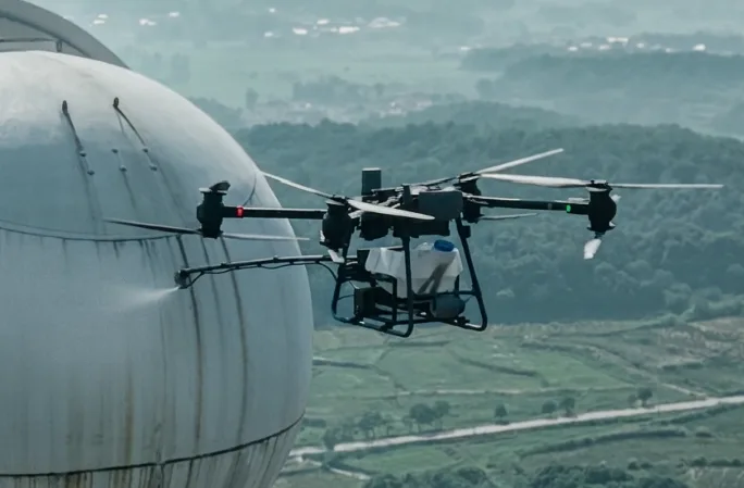 Aerial cleaning drone next to a curved tank wall above green hills