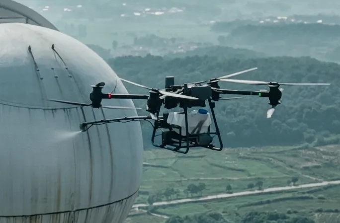 Drone with the cleaning system spraying a storage tank above green hills